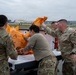 A group of U.S. Air Force personnel in OCP (Operational Camouflage Pattern) uniforms are unloading or loading large orange bags from the bed of a white pickup truck on what appears to be a military airfield. The orange bags are branded with text including 'Storage Chamber' and 'Princess Cruise' logos, suggesting these may be emergency or survival equipment items. The activity appears to be a coordinated logistics or equipment distribution effort on the flight line.