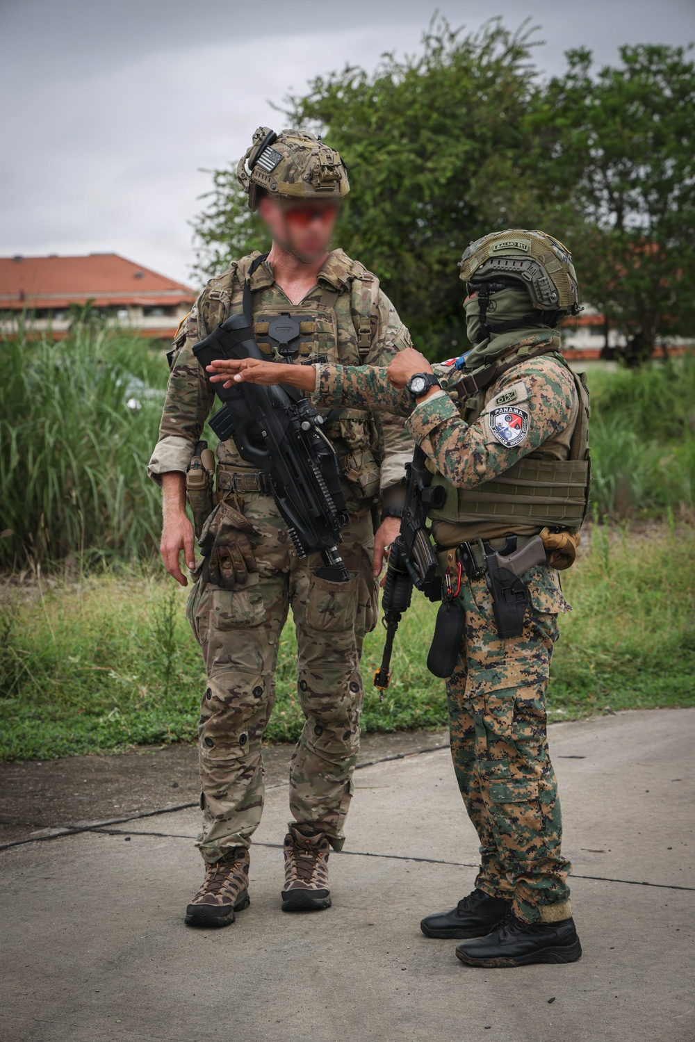 Two armed military personnel are standing on a paved surface in what appears to be a training or operational environment. One individual wears MultiCam/OCP pattern with a tactical helmet and carries a compact assault rifle, while the other wears a digital woodland camouflage pattern with a patch reading 'PANAMA' visible on the chest rig. The two appear to be engaged in a briefing or equipment inspection interaction.
