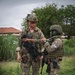 Two armed military personnel are standing on a paved surface in what appears to be a training or operational environment. One individual wears MultiCam/OCP pattern with a tactical helmet and carries a compact assault rifle, while the other wears a digital woodland camouflage pattern with a patch reading 'PANAMA' visible on the chest rig. The two appear to be engaged in a briefing or equipment inspection interaction.