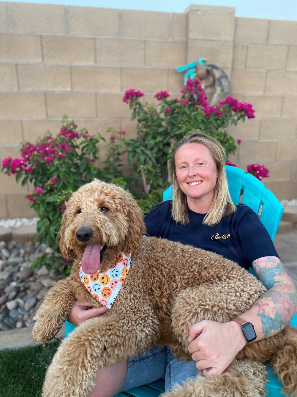 A smiling blonde woman sits in a turquoise Adirondack chair in a backyard, holding a large golden/apricot Goldendoodle dog wearing a smiley-face patterned bandana. The setting features a cinder block wall, blooming bougainvillea with bright pink flowers, and decorative yard elements. The woman is wearing a navy Pendleton t-shirt, jeans, and has visible tattoos on her right arm.