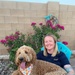 A smiling blonde woman sits in a turquoise Adirondack chair in a backyard, holding a large golden/apricot Goldendoodle dog wearing a smiley-face patterned bandana. The setting features a cinder block wall, blooming bougainvillea with bright pink flowers, and decorative yard elements. The woman is wearing a navy Pendleton t-shirt, jeans, and has visible tattoos on her right arm.
