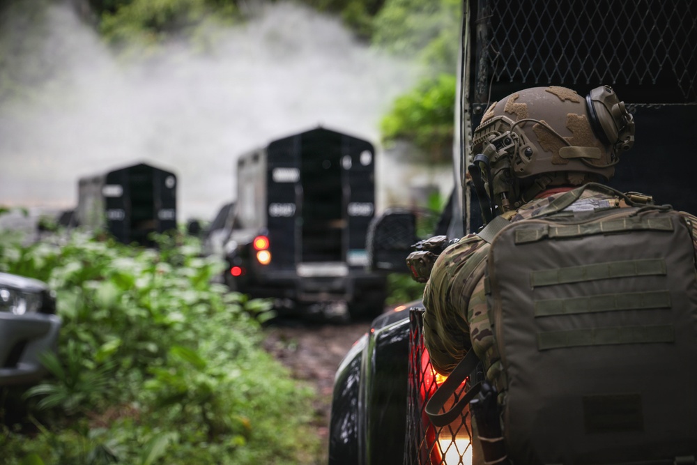 A tactical operator in full combat gear observes a convoy of armored vehicles moving along a narrow, vegetation-lined path. The scene depicts what appears to be a military or law enforcement operation in a jungle or heavily forested environment. Smoke or mist is visible in the background, suggesting recent activity or environmental conditions.