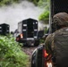 A tactical operator in full combat gear observes a convoy of armored vehicles moving along a narrow, vegetation-lined path. The scene depicts what appears to be a military or law enforcement operation in a jungle or heavily forested environment. Smoke or mist is visible in the background, suggesting recent activity or environmental conditions.