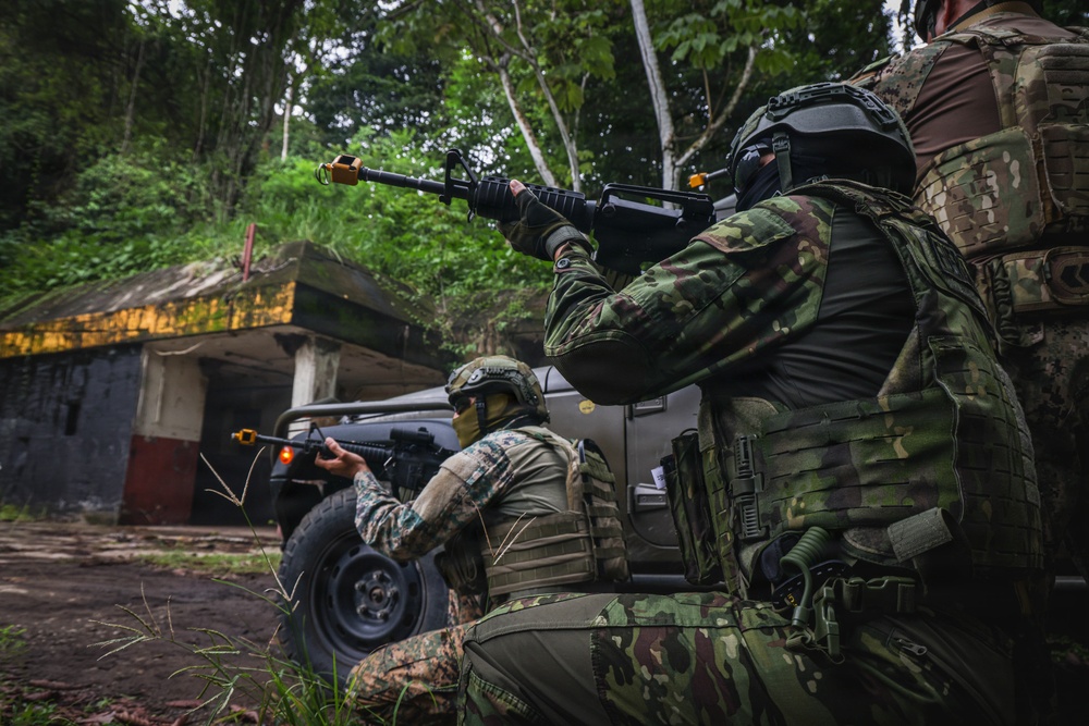 The image shows multiple armed personnel in military/tactical gear taking covered positions near a military-style vehicle in a jungle or forested environment. The personnel are equipped with rifles featuring orange muzzle devices, tactical vests, helmets, and face coverings. The setting includes a concrete structure in the background, suggesting a training exercise or tactical operation scenario.