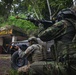 The image shows multiple armed personnel in military/tactical gear taking covered positions near a military-style vehicle in a jungle or forested environment. The personnel are equipped with rifles featuring orange muzzle devices, tactical vests, helmets, and face coverings. The setting includes a concrete structure in the background, suggesting a training exercise or tactical operation scenario.