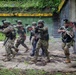 The image depicts a military or law enforcement tactical operation, likely a training exercise or live raid, in an outdoor urban-decay environment with tropical vegetation. Multiple armed personnel in varied camouflage uniforms are escorting what appears to be a detainee or role-player. At least one patch visible on a personnel member appears to be Peruvian, suggesting a multinational or joint operation context.