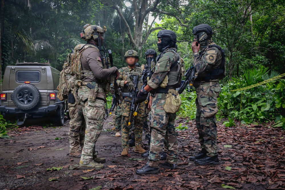 A group of approximately five heavily armed military or law enforcement personnel are gathered in a jungle or tropical forest clearing, appearing to conduct a briefing or coordination meeting. The personnel wear mixed camouflage patterns and tactical gear, suggesting a multinational or joint operation. A military-style vehicle is visible in the background, and yellow caution tape can be seen on the right side of the frame.
