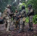 A group of approximately five heavily armed military or law enforcement personnel are gathered in a jungle or tropical forest clearing, appearing to conduct a briefing or coordination meeting. The personnel wear mixed camouflage patterns and tactical gear, suggesting a multinational or joint operation. A military-style vehicle is visible in the background, and yellow caution tape can be seen on the right side of the frame.