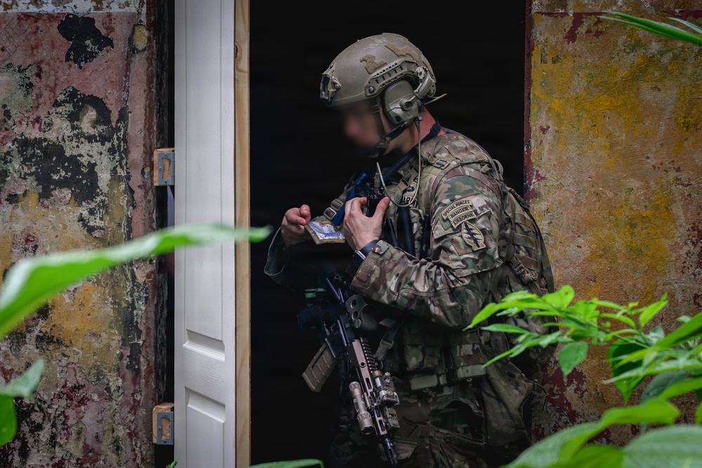 A U.S. Army Special Forces soldier in full tactical gear is positioned at a doorway of a weathered building, appearing to conduct a building entry or clearance operation. The soldier wears multicam camouflage with visible unit patches including Special Forces, Airborne, and a flash patch, and carries a rifle slung at low ready. The face has been deliberately blurred, consistent with operational security practices in official military imagery.