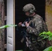 A U.S. Army Special Forces soldier in full tactical gear is positioned at a doorway of a weathered building, appearing to conduct a building entry or clearance operation. The soldier wears multicam camouflage with visible unit patches including Special Forces, Airborne, and a flash patch, and carries a rifle slung at low ready. The face has been deliberately blurred, consistent with operational security practices in official military imagery.