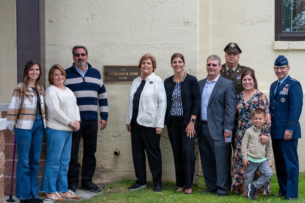 A group of approximately nine adults and one child pose for a formal photograph in front of a building plaque reading 'The Captain David M. Grimm Building.' The group includes civilians and two military personnel in uniform — one in Army dress uniform and one in Air Force dress uniform with a service cap. This appears to be a building dedication or naming ceremony.