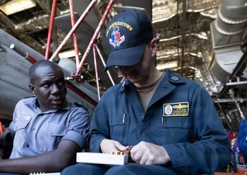 Two military personnel are seated in what appears to be a ship's hangar bay, with one sailor in dark blue coveralls loading ammunition into a magazine while another in gray uniform observes. The sailor in coveralls wears a cap reading 'USS Cooperstown' and a name tape patch reading 'L. Hofland, Operations.' Military aircraft components and ship infrastructure are visible in the background.