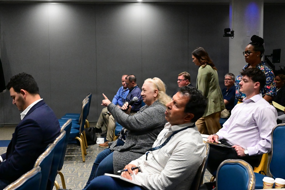 The image shows an indoor conference or seminar setting with approximately 10-12 attendees seated in blue and gold chairs. A woman with blonde hair is holding a microphone and pointing, suggesting she is asking a question or making a comment during a Q&amp;A session. Two women are standing in the background, possibly facilitating or observing the event.
