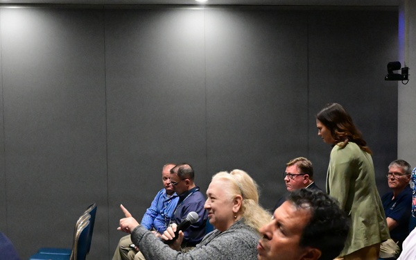 The image shows an indoor conference or seminar setting with approximately 10-12 attendees seated in blue and gold chairs. A woman with blonde hair is holding a microphone and pointing, suggesting she is asking a question or making a comment during a Q&amp;A session. Two women are standing in the background, possibly facilitating or observing the event.