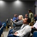 The image shows an indoor conference or seminar setting with approximately 10-12 attendees seated in blue and gold chairs. A woman with blonde hair is holding a microphone and pointing, suggesting she is asking a question or making a comment during a Q&amp;A session. Two women are standing in the background, possibly facilitating or observing the event.