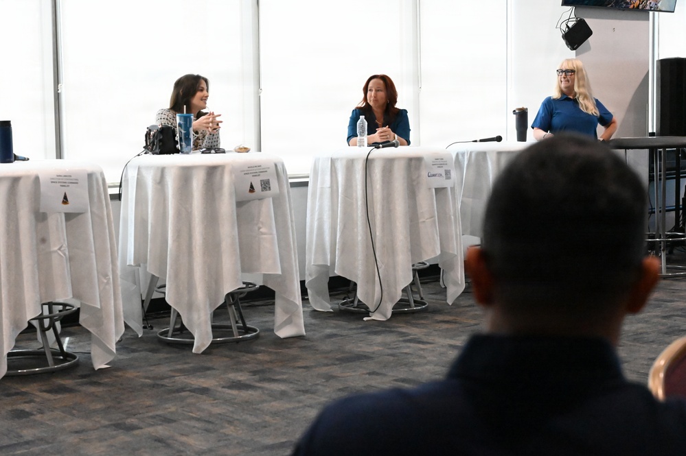 A panel discussion or forum is taking place in a modern conference or event space with large windows. Three women are seated at separate high-top tables draped in white tablecloths, with name placards visible in front of each. An audience member is visible in the foreground with their back to the camera.