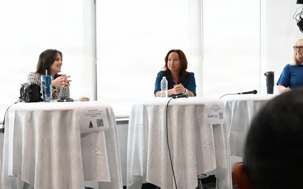 A panel discussion or forum is taking place in a modern conference or event space with large windows. Three women are seated at separate high-top tables draped in white tablecloths, with name placards visible in front of each. An audience member is visible in the foreground with their back to the camera.