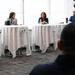 A panel discussion or forum is taking place in a modern conference or event space with large windows. Three women are seated at separate high-top tables draped in white tablecloths, with name placards visible in front of each. An audience member is visible in the foreground with their back to the camera.