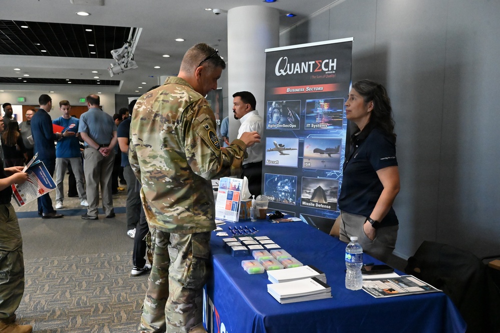 A military career fair or defense industry expo is taking place in a modern conference facility. A soldier in Army Combat Uniform (ACU) is engaging with a representative at the Quantech Services Inc. booth, which highlights business sectors including Agile DevSecOps, IT Systems, Aircraft, ISR, C-SISR, and Missile Defense. Multiple other attendees and booths are visible in the background.