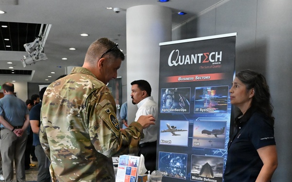 A military career fair or defense industry expo is taking place in a modern conference facility. A soldier in Army Combat Uniform (ACU) is engaging with a representative at the Quantech Services Inc. booth, which highlights business sectors including Agile DevSecOps, IT Systems, Aircraft, ISR, C-SISR, and Missile Defense. Multiple other attendees and booths are visible in the background.