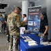 A military career fair or defense industry expo is taking place in a modern conference facility. A soldier in Army Combat Uniform (ACU) is engaging with a representative at the Quantech Services Inc. booth, which highlights business sectors including Agile DevSecOps, IT Systems, Aircraft, ISR, C-SISR, and Missile Defense. Multiple other attendees and booths are visible in the background.