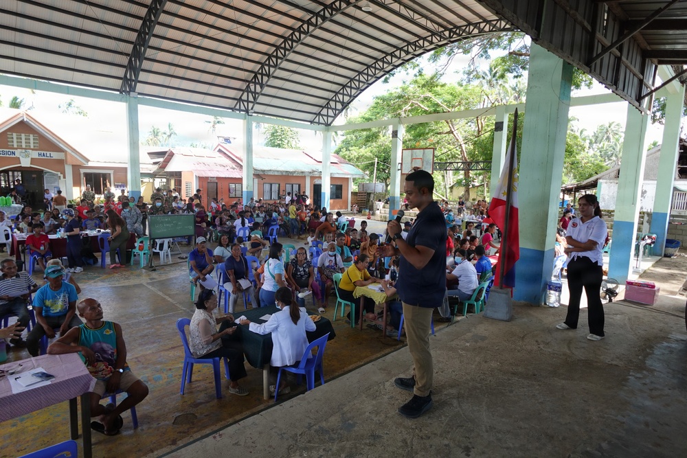 A large community gathering is taking place under an open-air covered pavilion, likely in the Philippines as indicated by the visible Philippine flag. The event appears to be a medical or social services outreach mission, with healthcare workers in white coats attending to seated community members. A male speaker is addressing the crowd using a microphone while a large number of residents are seated in plastic chairs throughout the venue.