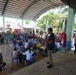A large community gathering is taking place under an open-air covered pavilion, likely in the Philippines as indicated by the visible Philippine flag. The event appears to be a medical or social services outreach mission, with healthcare workers in white coats attending to seated community members. A male speaker is addressing the crowd using a microphone while a large number of residents are seated in plastic chairs throughout the venue.