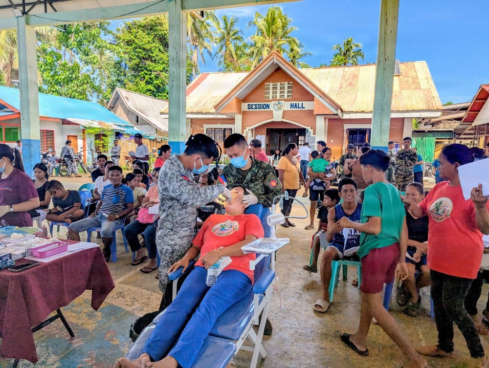 A military-organized medical/dental outreach mission is taking place in an open-air covered area adjacent to a 'Session Hall' building, likely in the Philippines. Military personnel in camouflage uniforms are providing dental treatment to civilians, with a patient reclined in a portable dental chair receiving oral care. A large crowd of community residents, including children and adults, are waiting their turn or observing the proceedings.
