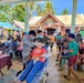 A military-organized medical/dental outreach mission is taking place in an open-air covered area adjacent to a 'Session Hall' building, likely in the Philippines. Military personnel in camouflage uniforms are providing dental treatment to civilians, with a patient reclined in a portable dental chair receiving oral care. A large crowd of community residents, including children and adults, are waiting their turn or observing the proceedings.