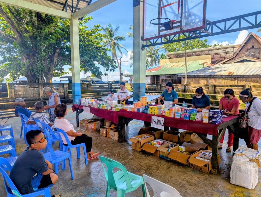 A community pharmacy or medical outreach event is taking place under an open-air covered basketball court. Healthcare workers wearing masks are stationed behind a long table labeled 'PHARMACY,' distributing medications and supplies to community members seated in plastic chairs. The setting appears to be in a rural or semi-urban area in the Philippines, based on the architecture, vegetation, and signage.