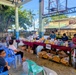 A community pharmacy or medical outreach event is taking place under an open-air covered basketball court. Healthcare workers wearing masks are stationed behind a long table labeled 'PHARMACY,' distributing medications and supplies to community members seated in plastic chairs. The setting appears to be in a rural or semi-urban area in the Philippines, based on the architecture, vegetation, and signage.