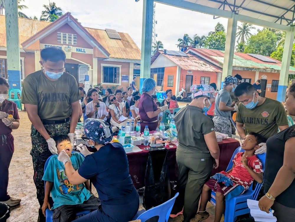A military medical/dental outreach mission is being conducted outdoors in what appears to be a Philippine barangay, with personnel wearing 'MARINES' uniforms providing dental care to children and community members. Multiple medical personnel in military attire and scrubs are treating patients at tables set up under a covered area. A building labeled 'SESSION HALL' is visible in the background, indicating this is likely a local government facility being used as a staging area.