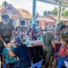 A military medical/dental outreach mission is being conducted outdoors in what appears to be a Philippine barangay, with personnel wearing 'MARINES' uniforms providing dental care to children and community members. Multiple medical personnel in military attire and scrubs are treating patients at tables set up under a covered area. A building labeled 'SESSION HALL' is visible in the background, indicating this is likely a local government facility being used as a staging area.