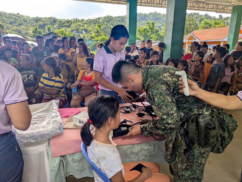 A military medical outreach or civil-military operations event is taking place in an open-air community structure, likely in a rural Southeast Asian (possibly Philippine) setting. A soldier in camouflage uniform is using a stethoscope to examine or take the blood pressure of a young girl seated at a table, assisted by a civilian health worker. A large crowd of community residents, including children and adults, are waiting in the background for their turn.