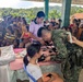 A military medical outreach or civil-military operations event is taking place in an open-air community structure, likely in a rural Southeast Asian (possibly Philippine) setting. A soldier in camouflage uniform is using a stethoscope to examine or take the blood pressure of a young girl seated at a table, assisted by a civilian health worker. A large crowd of community residents, including children and adults, are waiting in the background for their turn.