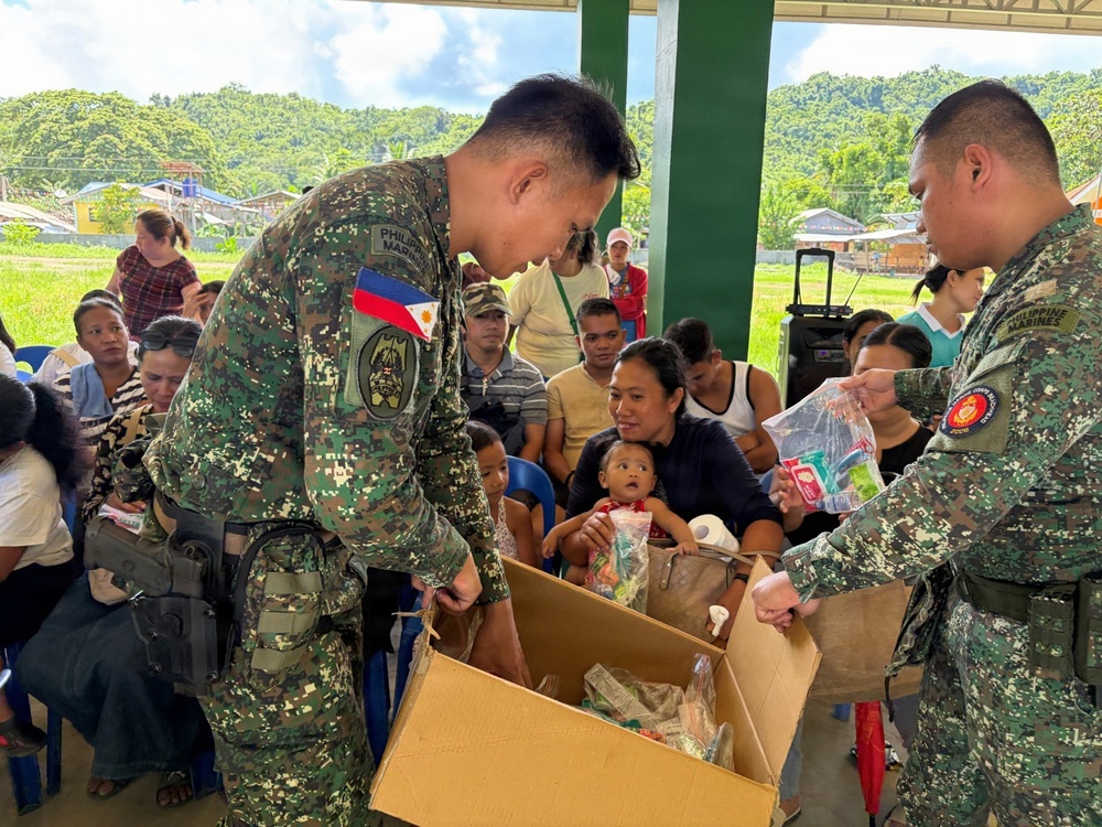 Two Philippine Marines in digital camouflage uniforms are distributing relief goods from a cardboard box to civilians gathered under an open-air covered structure. A crowd of community members, including women and children, are seated waiting to receive assistance. The setting appears to be a rural or semi-rural area in the Philippines, consistent with a humanitarian aid or civil-military operations activity.
