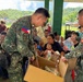 Two Philippine Marines in digital camouflage uniforms are distributing relief goods from a cardboard box to civilians gathered under an open-air covered structure. A crowd of community members, including women and children, are seated waiting to receive assistance. The setting appears to be a rural or semi-rural area in the Philippines, consistent with a humanitarian aid or civil-military operations activity.