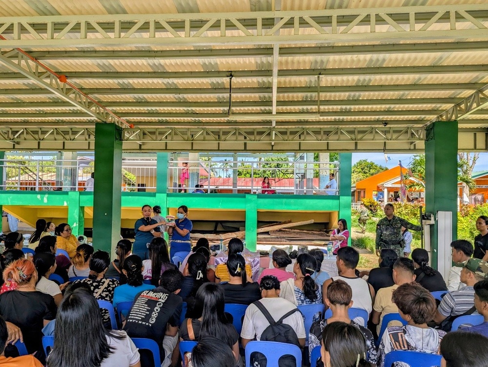 A community gathering or outreach event is taking place under an open-air covered pavilion, likely in the Philippines based on architectural style and vegetation. A large crowd of civilians seated on blue plastic chairs faces two presenters or facilitators at the front, with at least one uniformed military or law enforcement personnel visible on the right side. The setting appears to be a barangay hall or school compound with colorful green and orange buildings visible in the background.