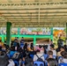A community gathering or outreach event is taking place under an open-air covered pavilion, likely in the Philippines based on architectural style and vegetation. A large crowd of civilians seated on blue plastic chairs faces two presenters or facilitators at the front, with at least one uniformed military or law enforcement personnel visible on the right side. The setting appears to be a barangay hall or school compound with colorful green and orange buildings visible in the background.