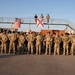 A large formation of military personnel in OCP (Operational Camouflage Pattern) uniforms stand at parade rest facing an elevated platform structure. Two flags are displayed on the platform: the United States flag and the Florida state flag, suggesting this is a Florida National Guard unit. The setting appears to be a military installation or forward operating base, likely during a formation ceremony or briefing.