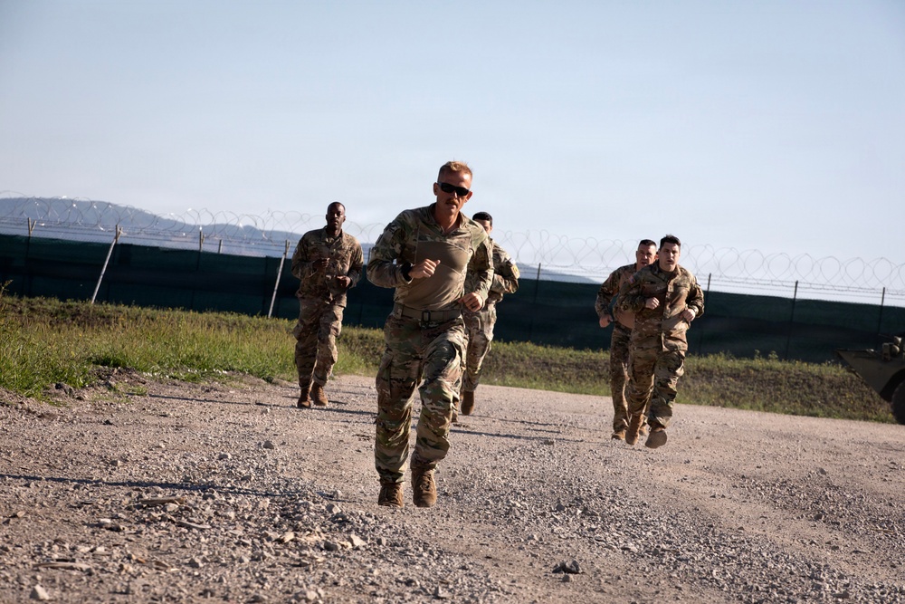 A group of military personnel in camouflage uniforms are running along a gravel path near a perimeter fence topped with razor wire. The lead soldier, wearing sunglasses, appears to be setting the pace for the group behind him. The setting appears to be a military installation or forward operating base, with hills visible in the background.