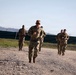 A group of military personnel in camouflage uniforms are running along a gravel path near a perimeter fence topped with razor wire. The lead soldier, wearing sunglasses, appears to be setting the pace for the group behind him. The setting appears to be a military installation or forward operating base, with hills visible in the background.