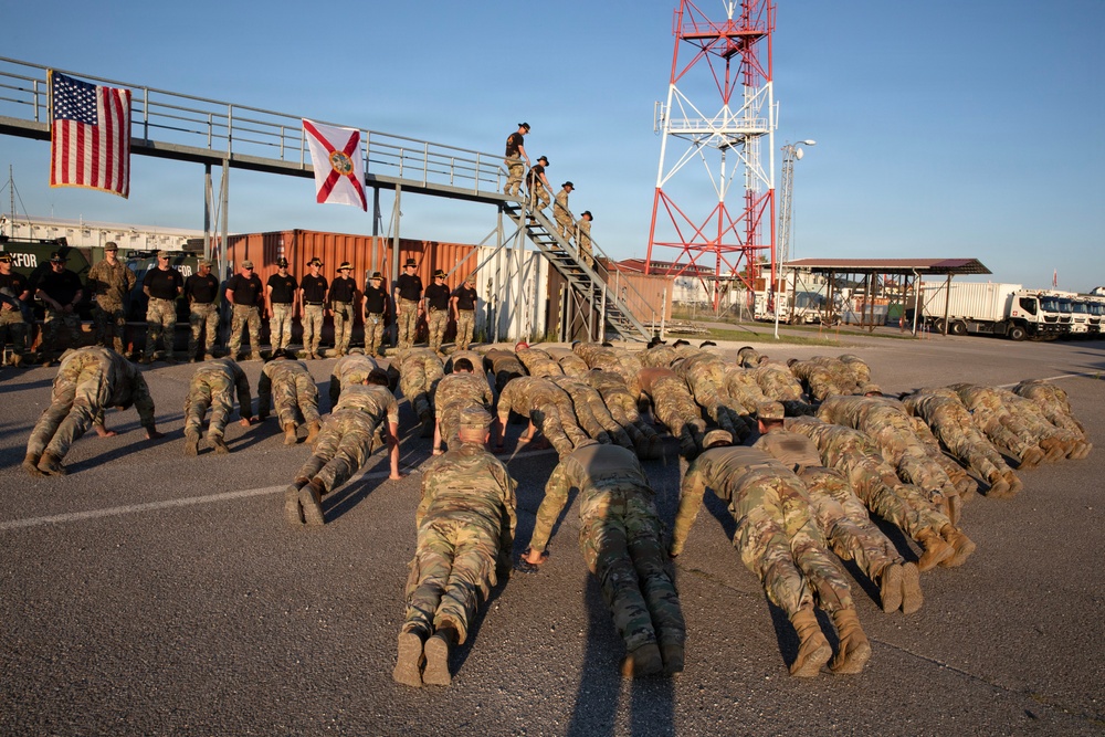 A large group of military personnel in camouflage uniforms are performing push-ups on a paved surface during what appears to be a physical training exercise or ceremony. A separate group of soldiers in matching uniforms stands at attention observing the exercise, with an American flag and what appears to be the Florida state flag displayed on a structure behind them. The setting appears to be a military installation or port facility with shipping containers, vehicles, and a red-and-white communications tower visible.