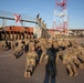 A large group of military personnel in camouflage uniforms are performing push-ups on a paved surface during what appears to be a physical training exercise or ceremony. A separate group of soldiers in matching uniforms stands at attention observing the exercise, with an American flag and what appears to be the Florida state flag displayed on a structure behind them. The setting appears to be a military installation or port facility with shipping containers, vehicles, and a red-and-white communications tower visible.