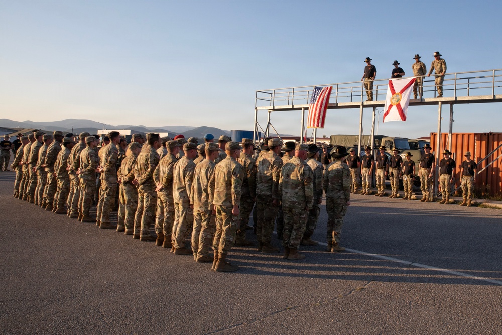 A large formation of military personnel in camouflage uniforms are assembled on a paved surface, likely during a ceremony or formal muster. Two flags — the United States flag and what appears to be the Florida state flag — are displayed on an elevated platform structure where four individuals in civilian or instructor attire are standing. The setting appears to be an outdoor military installation or training facility at either dawn or dusk based on the warm, low-angle lighting.