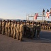 A large formation of military personnel in camouflage uniforms are assembled on a paved surface, likely during a ceremony or formal muster. Two flags — the United States flag and what appears to be the Florida state flag — are displayed on an elevated platform structure where four individuals in civilian or instructor attire are standing. The setting appears to be an outdoor military installation or training facility at either dawn or dusk based on the warm, low-angle lighting.