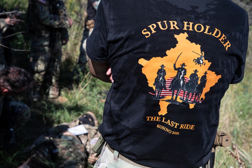 A person wearing a black t-shirt with the text 'SPUR HOLDER - THE LAST RIDE - KOSOVO 2026' is shown from behind. The shirt features an orange graphic depicting horseback riders with a military helicopter against a silhouette of Kosovo's map. The setting appears to be an outdoor military training or exercise environment with other personnel in camouflage uniforms visible in the background.