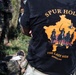 A person wearing a black t-shirt with the text 'SPUR HOLDER - THE LAST RIDE - KOSOVO 2026' is shown from behind. The shirt features an orange graphic depicting horseback riders with a military helicopter against a silhouette of Kosovo's map. The setting appears to be an outdoor military training or exercise environment with other personnel in camouflage uniforms visible in the background.