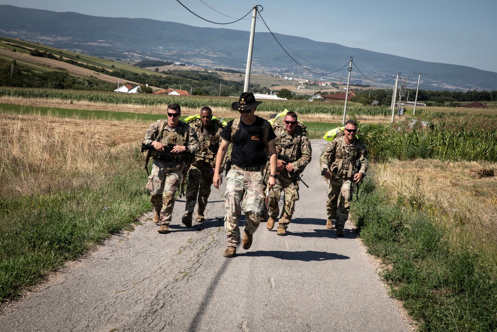 A group of five military personnel in combat gear are marching along a narrow rural road through an agricultural landscape. The lead individual wears a distinctive black wide-brim hat and a black t-shirt with camouflage pants, while the others wear full military uniforms with tactical vests and backpacks. The setting appears to be a rural valley with cornfields, rolling hills, and scattered buildings visible in the background.