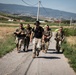 A group of five military personnel in combat gear are marching along a narrow rural road through an agricultural landscape. The lead individual wears a distinctive black wide-brim hat and a black t-shirt with camouflage pants, while the others wear full military uniforms with tactical vests and backpacks. The setting appears to be a rural valley with cornfields, rolling hills, and scattered buildings visible in the background.